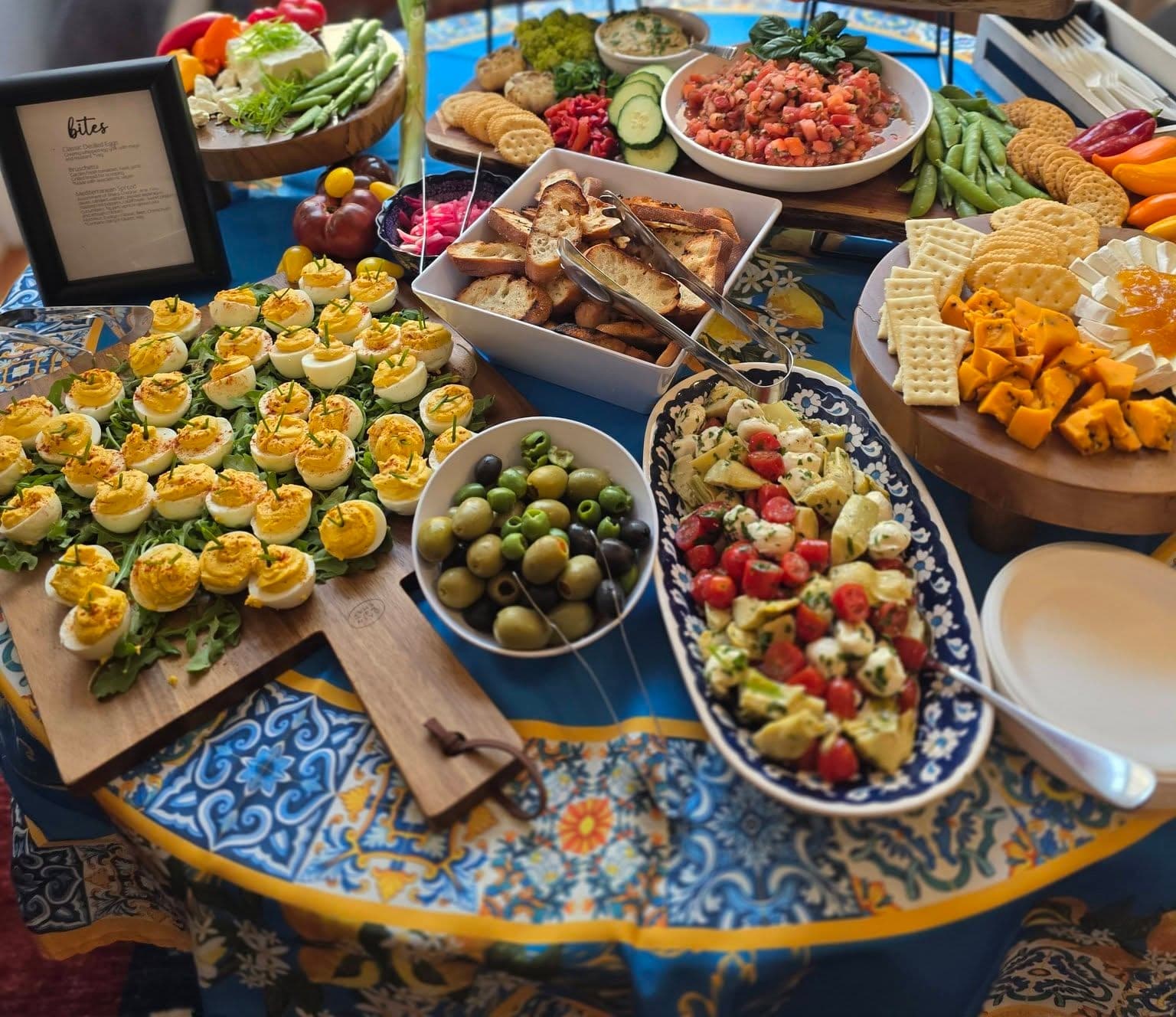 A round table on a blue and yellow patterned tablecloth, set with deviled eggs on arugula, olives, a tomato, mozzarella and artichoke salad, toasted bread, crackers and cheeses, beside a framed menu card.